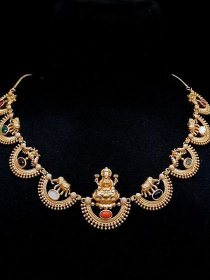 This unique necklace features a central Lakshmi motif with elephant figures and navratna stones set in half-moon shapes. It's a very distinctive piece of temple jewellery.
