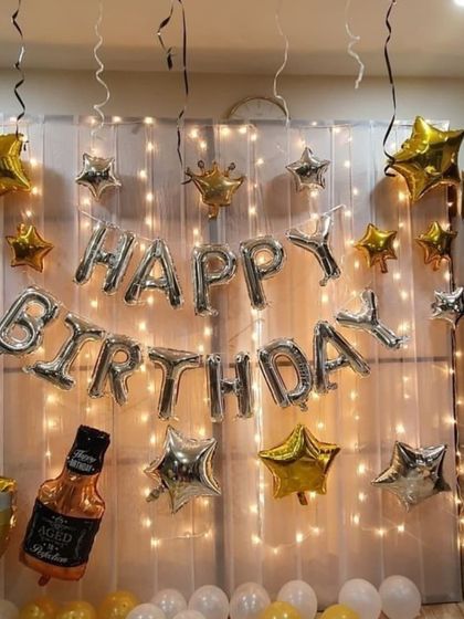 For this birthday setup, I used a wall of fairy lights behind a sheer white curtain to create a magical effect. The silver 'Happy Birthday' balloons are accented with gold and silver star balloons and a fun whiskey bottle prop.