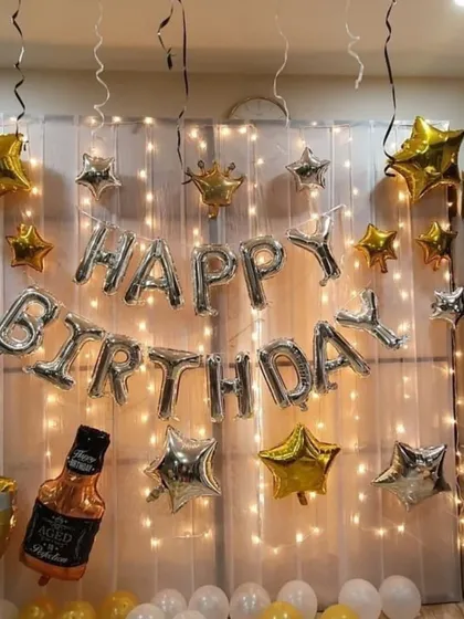 For this birthday setup, I used a wall of fairy lights behind a sheer white curtain to create a magical effect. The silver 'Happy Birthday' balloons are accented with gold and silver star balloons and a fun whiskey bottle prop.