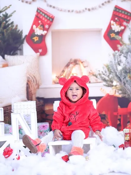 A cozy Christmas scene with a baby in a cute red hooded jacket, sitting among presents and festive decorations.