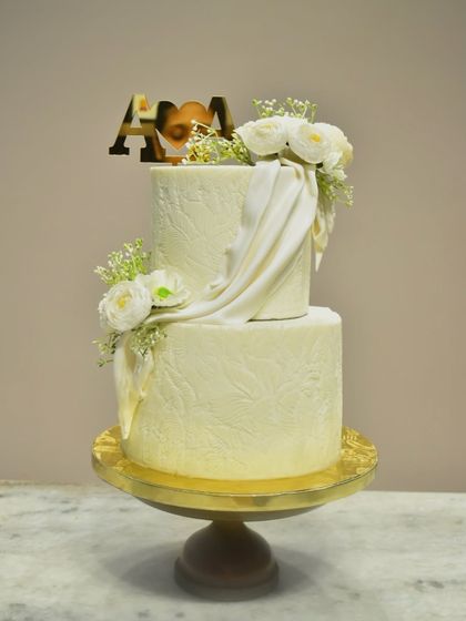 An elegant two-tier engagement cake with a subtle, embossed leaf pattern on the buttercream. The design is elevated with a flowing white fondant drape and fresh white flowers, complemented by a custom gold initial topper.