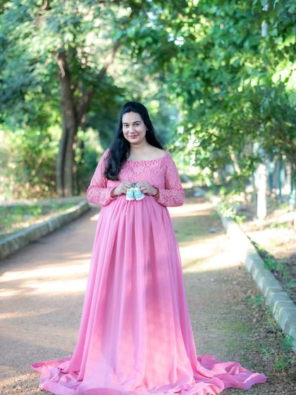 A classic outdoor portrait of a mom-to-be in a pink gown, holding a pair of tiny baby shoes on a park path.