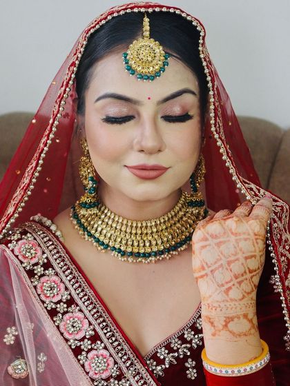 A close-up of a bride in a deep red velvet lehenga, with soft glam makeup and green and gold jewellery.