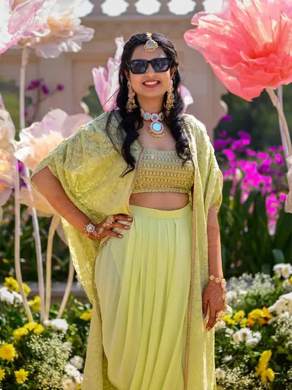 A bride looking chic and happy at her Mehendi ceremony. She wears a custom lime green outfit with a crop top, draped pants, and an embroidered cape.