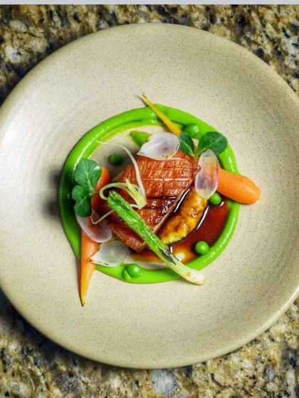 A beautifully plated main course. This dish features pan-seared duck breast on a bed of vibrant green pea puree, garnished with baby carrots, asparagus, and edible flowers for a fine-dining look.