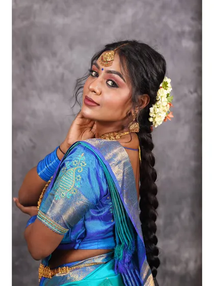 A profile shot showcasing the elegant drape of a blue silk saree and the traditional long braid. This angle is perfect for highlighting hair, makeup, and earrings.