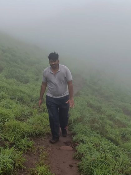 A trekker walking along a narrow path through the tall, wet grass on the way to Bandaje Falls, surrounded by thick fog.