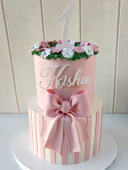 A dreamy two-tiered cake for a first birthday. The design features a pink and white striped bottom tier with a large bow, and a pink top tier with a floral crown and the number '1'.