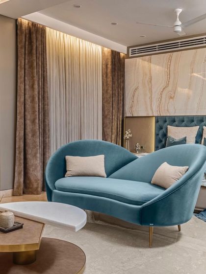 A curvilinear chaise lounge in a striking teal fabric adds a soft, sculptural element to the master bedroom. It provides a comfortable reading spot while complementing the room's luxurious material palette.