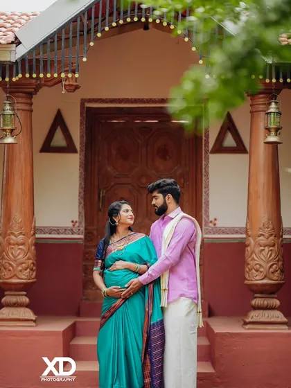 A loving couple's portrait in front of a traditional house, their hands together on the baby bump.