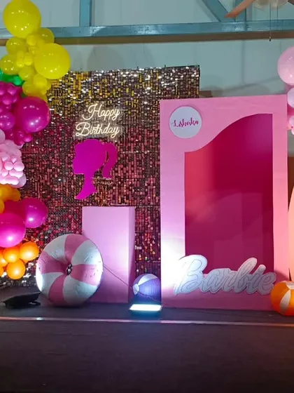 A vibrant, tropical Barbie-themed birthday party. This setup features a pink "doll box" photo booth, a sequin backdrop, and colorful balloon clusters with palm tree cutouts.