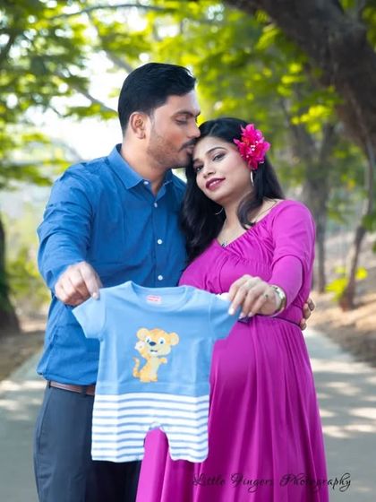 A sweet announcement photo where the couple holds up a tiny blue baby romper. The mother's vibrant pink dress and the natural park path create a lovely scene.
