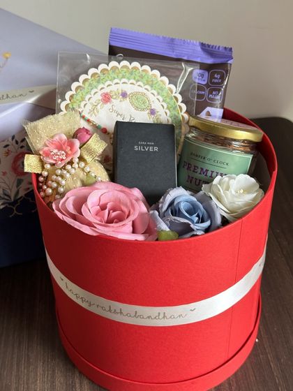 A compact and charming Rakhi hamper in a round red box. It includes a Zara mini perfume, a decorated coconut, a protein cookie, and a jar of dry fruits.