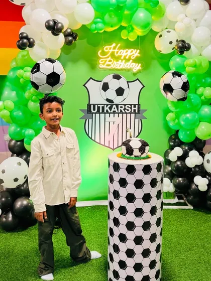 The birthday boy, Utkarsh, standing proudly in front of his football-themed birthday setup. The personalized shield and soccer ball cake on a matching plinth made his day complete.