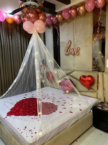 A simple yet romantic bed decoration. This setup includes a heart made of rose petals, a fairy light canopy, and foil balloons spelling 'love' on the headboard.