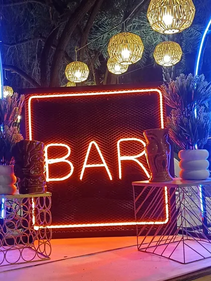 A rustic yet modern bar entrance design. The red neon 'BAR' sign is framed by tiki-style decor and woven hanging lamps, perfect for an outdoor or bohemian-themed event.