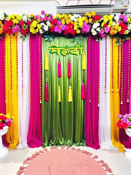 A vibrant Mehendi backdrop with a green center panel, flanked by pink and yellow drapes. The colorful tassels and floral arrangements add to the festive look.