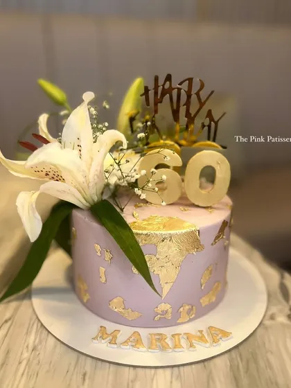 A chic lavender cake for Marina's 60th birthday. The design is elevated with gold leaf details, a "Happy 60" topper, and fresh white lilies.