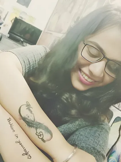 A client showing off her two forearm tattoos: a "Mom Dad" infinity feather design and a "Believe in Yourself" script tattoo.