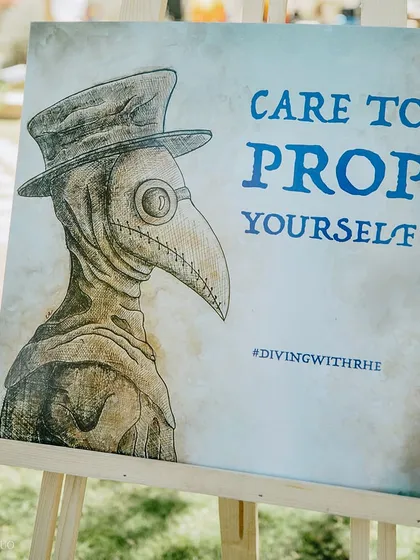 A quirky sign at the prop station for a festival-themed party. The "Care to Prop Yourself" sign, featuring a plague doctor illustration, invited guests to get creative with their look.