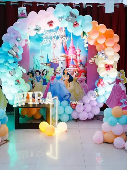 A magical Disney Princess theme for a first birthday. This enchanting setup features a pastel rainbow balloon arch, a castle backdrop, and life-sized cutouts of Cinderella, Snow White, and other princesses, creating a fairytale experience.