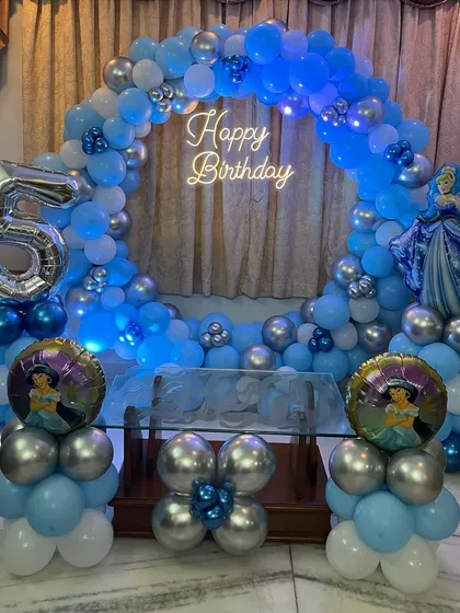 A Frozen themed home decoration featuring a circular balloon arch in shades of blue and silver, a neon sign, and foil balloons of Princess Jasmine, adding a unique twist.