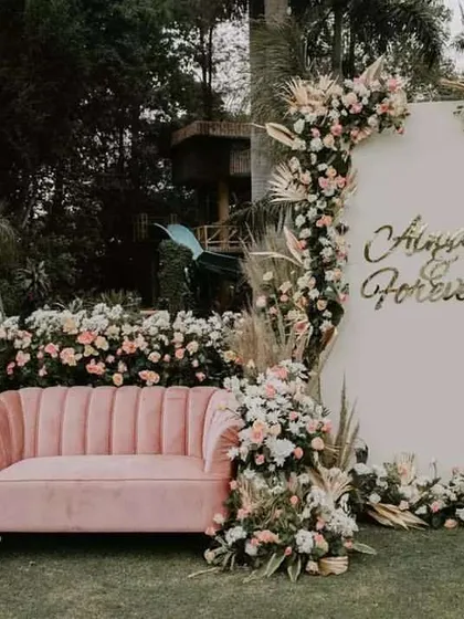 Here is a modern and chic photo booth for an outdoor event. It features a comfortable pink sofa, a stylish white backdrop with custom gold lettering, and a beautiful arrangement of pampas grass and pastel flowers for a bohemian touch.