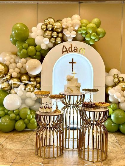 This First Holy Communion stage decoration uses a green, white, and gold color scheme. The setup includes an arched backdrop with a cross, a balloon garland, and stylish dessert stands.