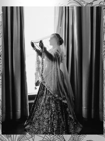 A beautiful silhouette of the bride against the window. This black and white shot captures the grandeur of her lehenga and the delicate transparency of her veil, creating a timeless and artistic bridal portrait.