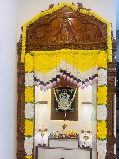 For housewarmings and festivals, I provide elegant pooja room decorations. This setup features intricate garlands of yellow and white flowers with lotus buds, designed to fit this beautiful carved wooden pooja mandir perfectly.