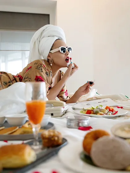 A playful "breakfast in bed" scene from a hotel lifestyle shoot. The model applies lipstick while surrounded by a luxurious breakfast spread, creating a fun and glamorous image.