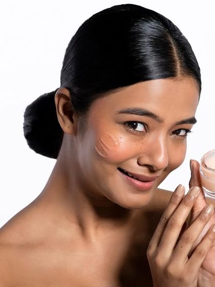 A friendly and inviting shot that showcases the product and its effect. The model's smile and direct gaze create a connection with the viewer, while the swatches on her cheek provide useful information about the product's appearance.