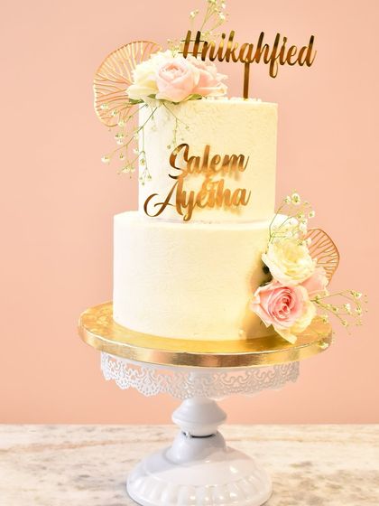 A two-tier cake designed for a Nikah ceremony, featuring custom gold acrylic toppers with the couple's names and "Nikahfied." The simple white buttercream is elegantly decorated with fresh roses and delicate gold fans.