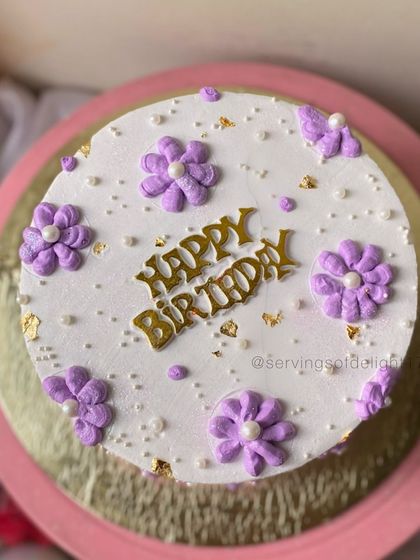 A top-down view of a simple birthday cake with piped purple flowers and gold leaf accents. It's a sweet and charming design.