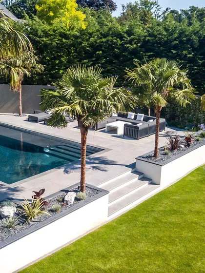 A luxurious poolside area featuring modern outdoor furniture and mature palm trees. We design elevated planters and use a mix of stone and lawn to create a sophisticated, multi-level resort feel.