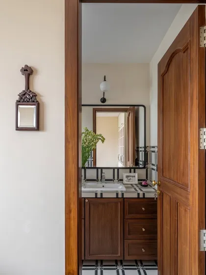 A peek through a wooden door reveals a bathroom that feels classic and elegant. The simple black-and-white tiles, dark wood vanity, and a small, carved mirror create a timeless look that is both understated and refined.