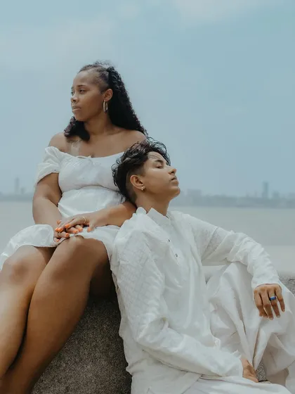 A serene and contemplative portrait of the couple sitting by the sea, with the Mumbai skyline in the distance. A quiet moment of togetherness.