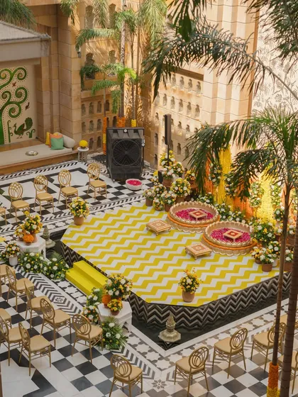 An aerial view of a modern Haldi setup in a royal courtyard. The design features a bold yellow and black chevron stage, traditional seating, and elegant floral arrangements.