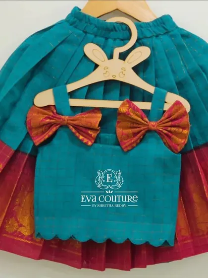 A modern and cute top and skirt set for a toddler. The teal top has adorable pattu bows on the shoulders, and the pleated skirt has a traditional pink and gold border.