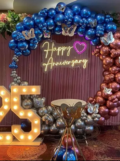 Celebrating a 25th anniversary with a modern color palette. This circular balloon arch combines metallic blue, chrome, and rose gold balloons, accented with butterfly details. The large light-up '25' and a 'Happy Anniversary' neon sign complete this stylish party decoration.