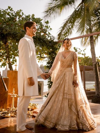 That just married feeling. The couple's coordinated ivory outfits look stunning in the golden hour light of their beach wedding. Her look is completed with a delicate veil and minimalist Purab Paschim jewelry.