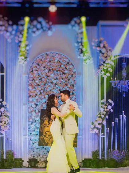 The couple's first dance at their elegant reception, set against a beautiful floral backdrop and dramatic stage lighting.