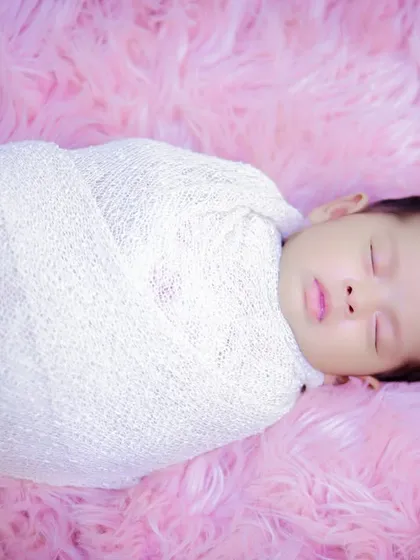 A beautifully simple shot of a newborn swaddled in white, sleeping on a soft, pink fur blanket. This highlights the baby's peaceful and angelic nature.