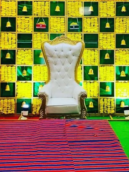 A vibrant and traditional stage for a naming ceremony. The backdrop is a bright green and yellow grid pattern decorated with small bells, with an ornate white throne chair as the focal point.