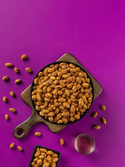 Masala-coated peanuts captured in a top-down composition. The deep purple background provides a rich contrast, highlighting the texture and color of the product for packaging.