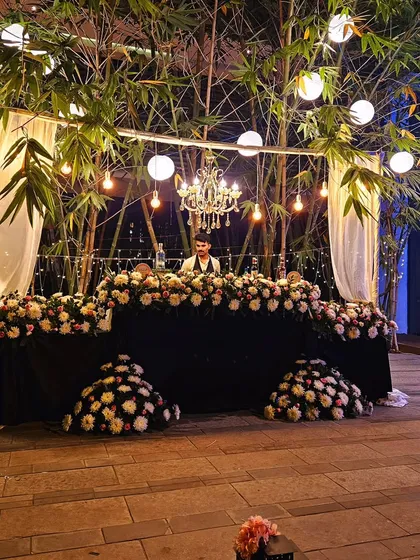 An elegant outdoor bar setup perfect for a sophisticated garden party. The bar is heavily decorated with white and pink flowers, set under a canopy of bamboo with hanging lanterns and a chandelier.
