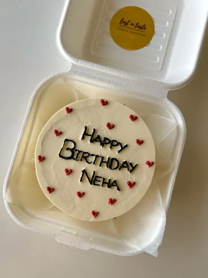 A classic birthday bento with a personal touch. This cake features "Happy Birthday Neha" in bold black icing, surrounded by small red hearts on a cream-colored base.