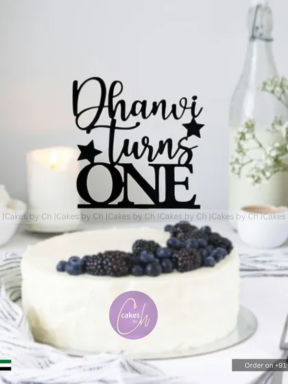 A mockup showing how the "Dhanvi turns ONE" topper in black acrylic would look on a simple white cake with berries.