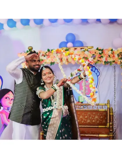 The couple poses with a floral bow and arrow in front of their baby shower backdrop, which features a 'It's a Girl' sign and balloon decorations.
