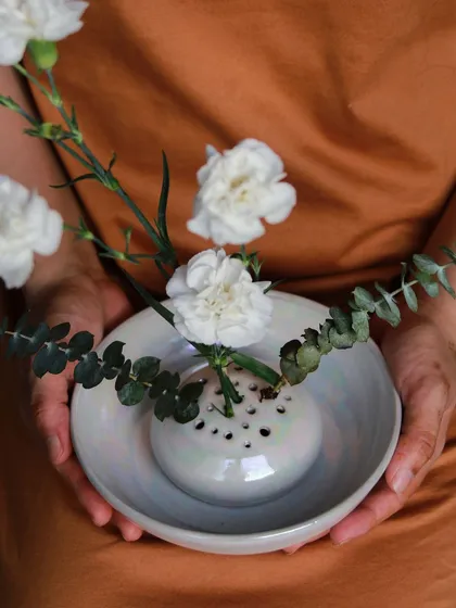 A delicate flower frog vase from the 'Celebrations' collection, held in hand to show its pearlescent glaze and intricate design.